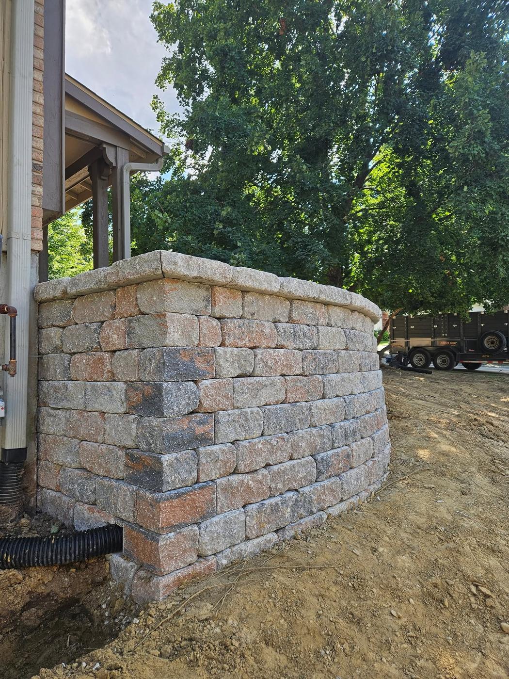 A curved block wall with capstones and a drain pipe at its base, fresh fill around it