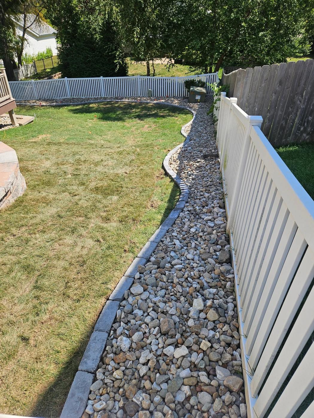 A river-rock drainage bed with edging running the length of a white fence