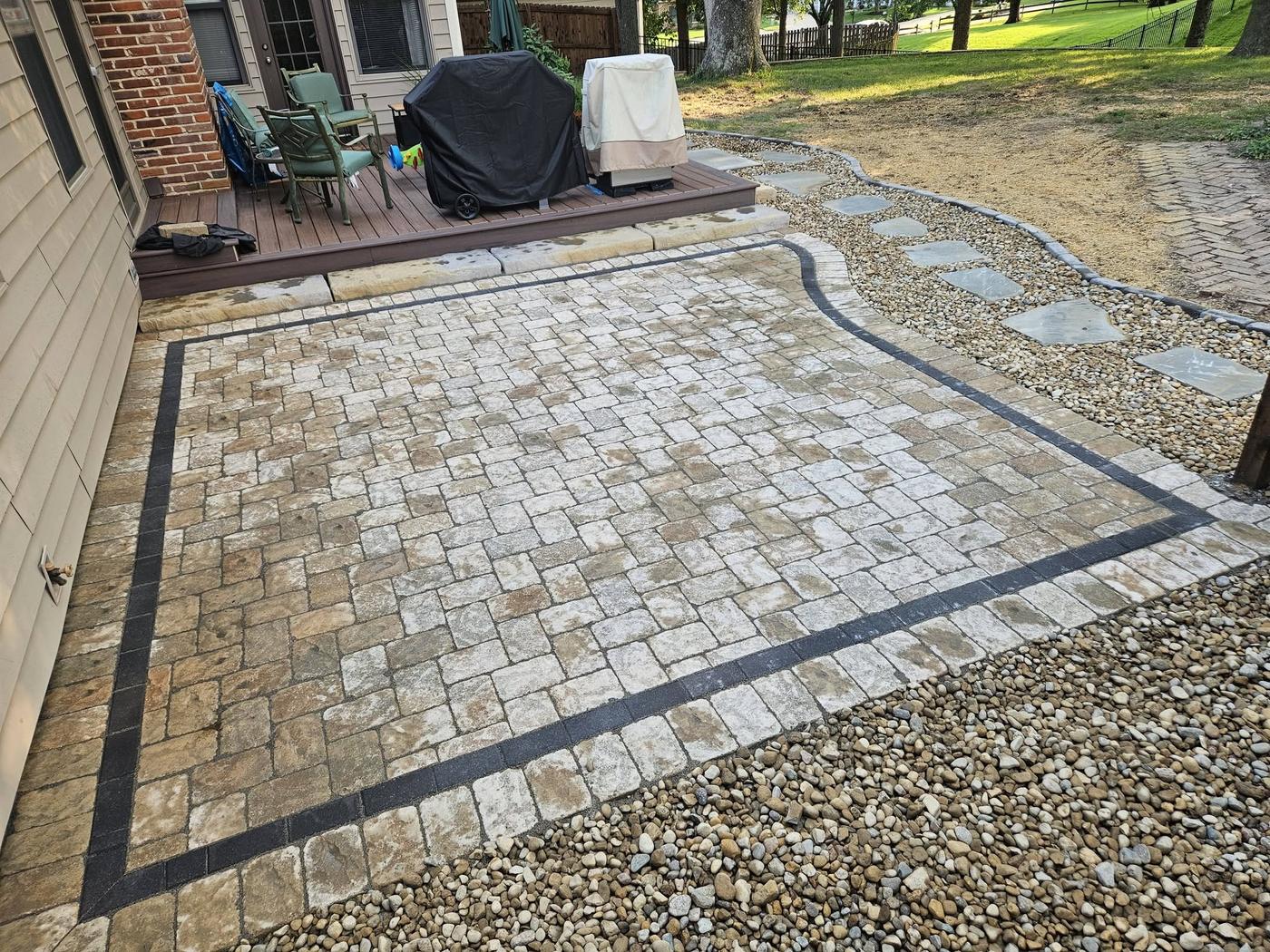 A rectangular paver patio with a dark border, gravel surround and steps up to a deck